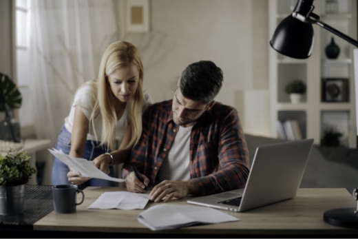 Zwei Personen arbeiten gemeinsam an einem Schreibtisch mit einem Laptop, Papieren und einer Tasse Kaffee.